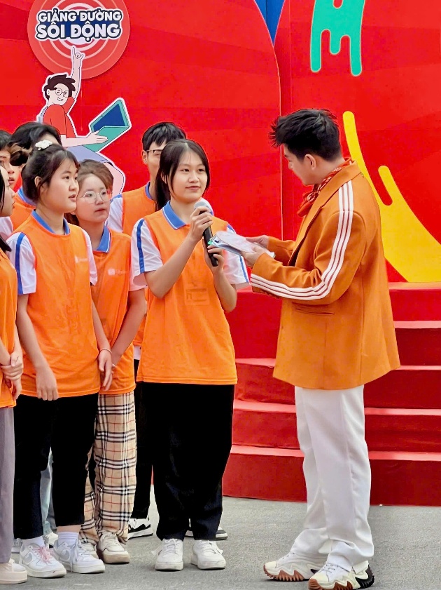 A person in orange shirt holding a microphone to a group of kids Description automatically generated