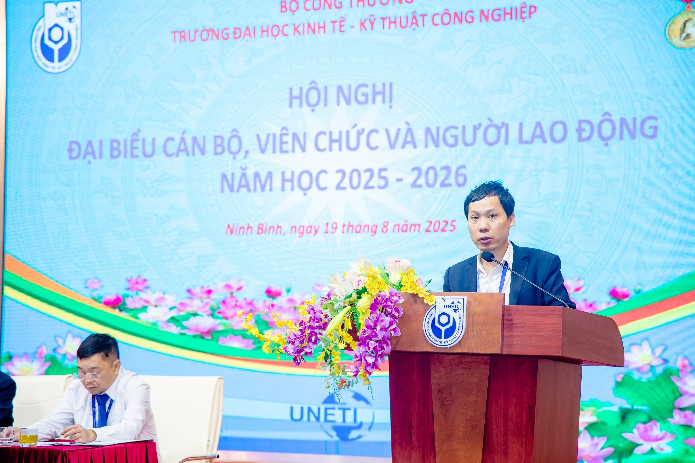 A person standing at a podium with a person behind him Description automatically generated