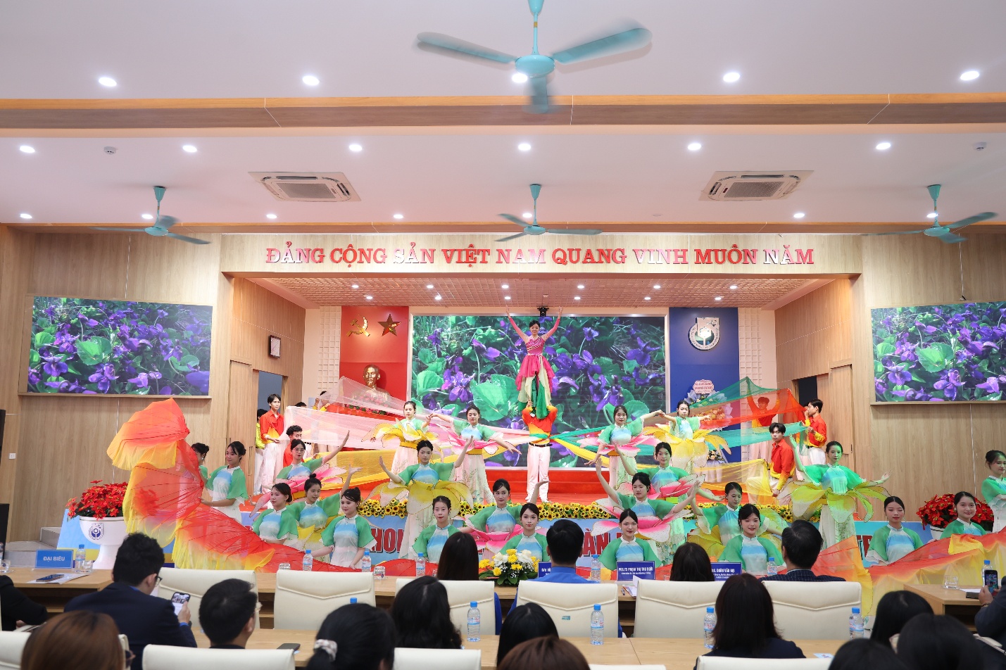 A group of people in a room with a stage and a fan Description automatically generated