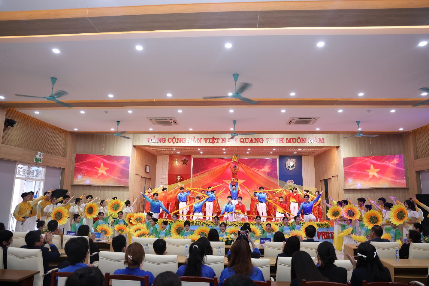 A group of people in a room with a stage and a group of people in the background Description automatically generated