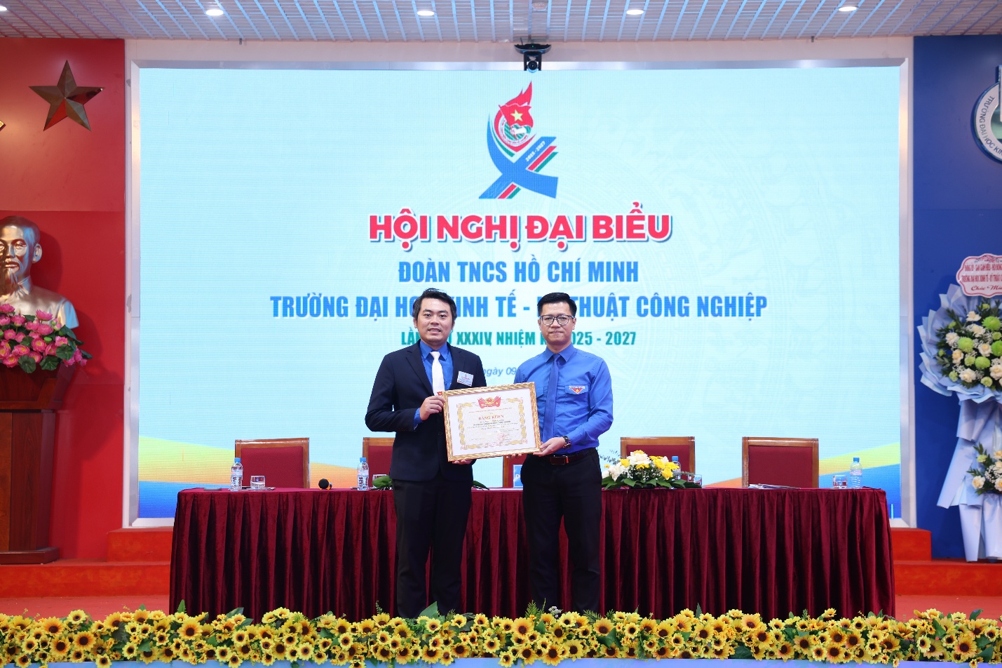 Two men standing on a stage holding a certificate Description automatically generated