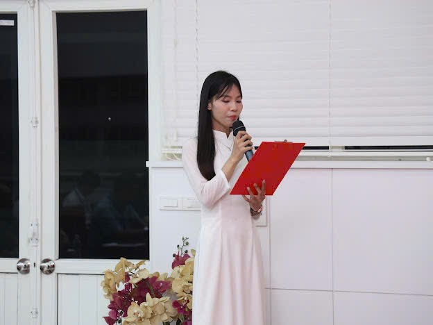 A person in a white dress holding a microphone and a red folder
Description automatically generated