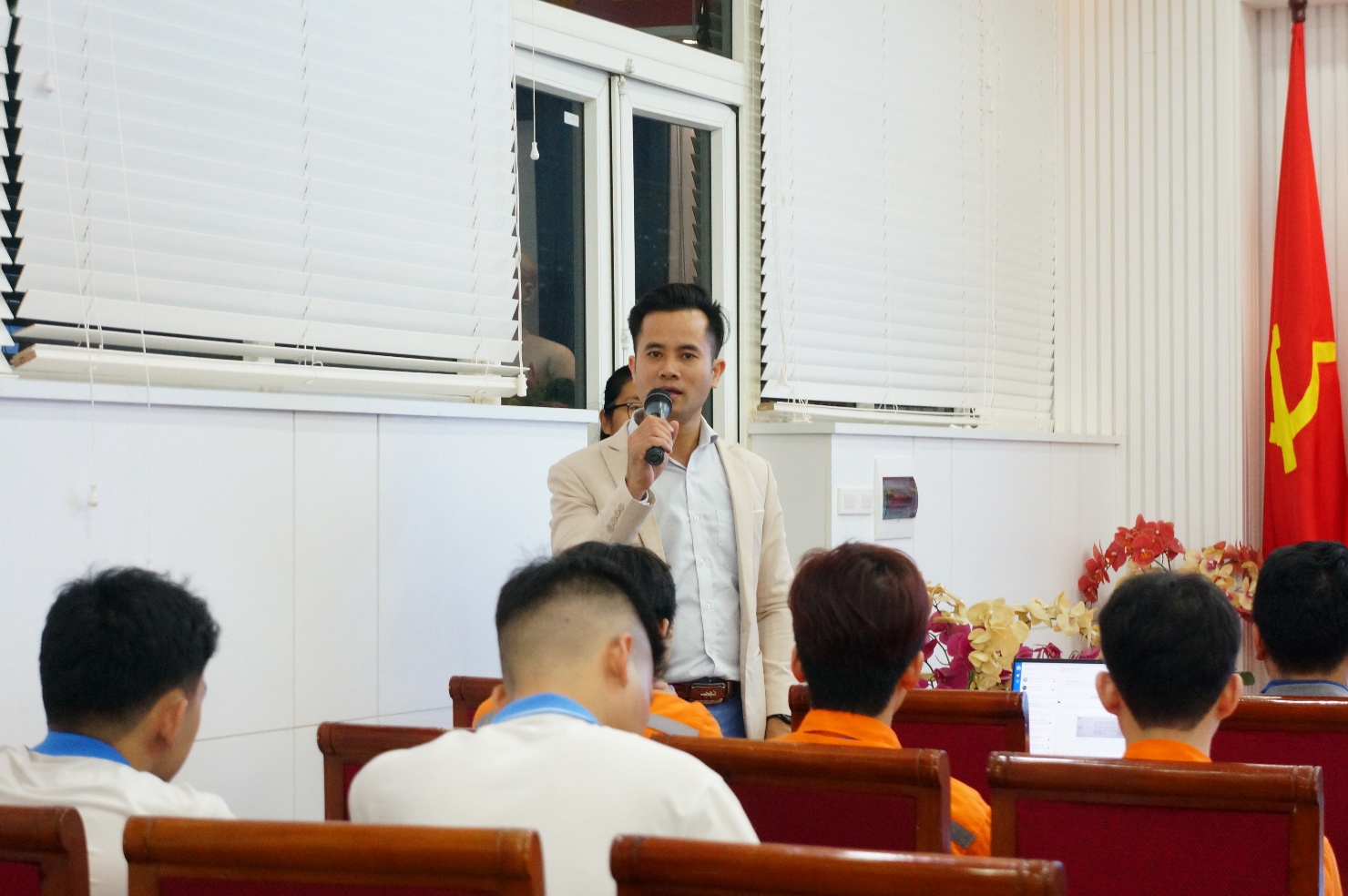 A person speaking into a microphone in front of a group of people Description automatically generated